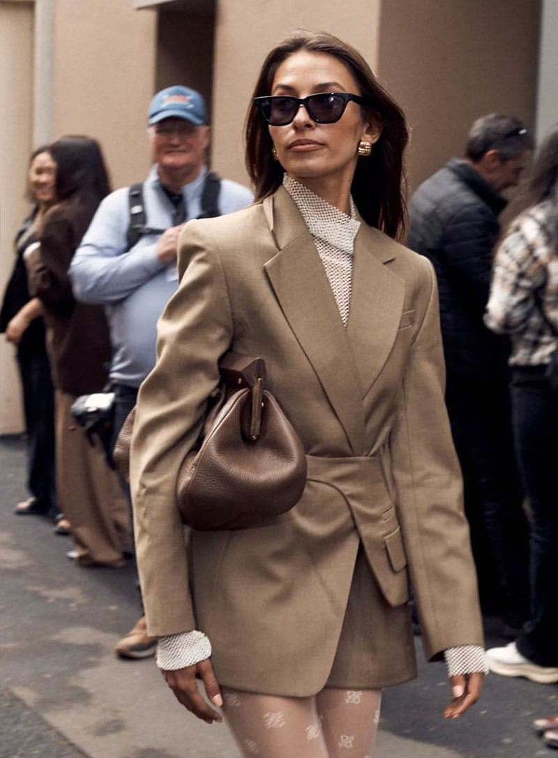 A woman walking outdoors wearing cat eye sunglassea and tailored beige blazer and matching mini skirt, sheer patterned tights, a white top, and carrying a brown leather bucket bag.