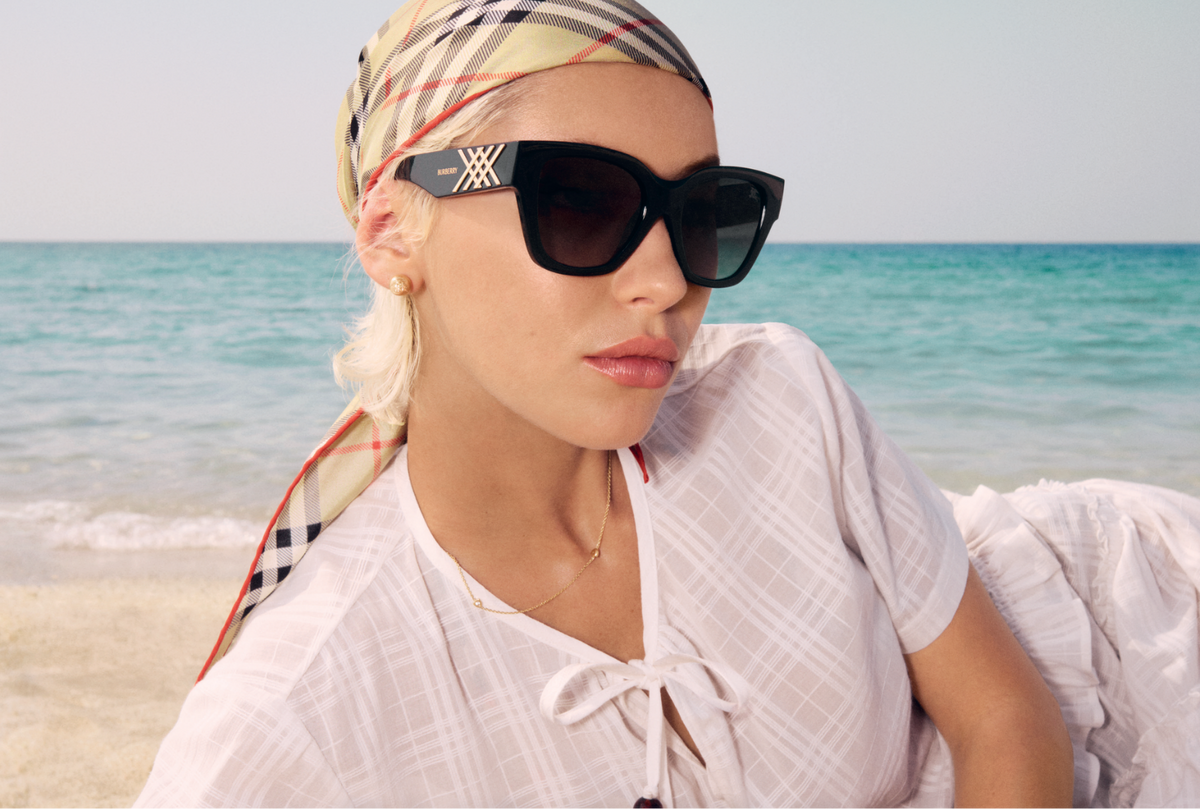 A woman on a beach wearing a Burberry plaid headscarf, black cat-eye sunglasses, and a white tie-front blouse. The word BURBERRY is printed in white across the lower center.
