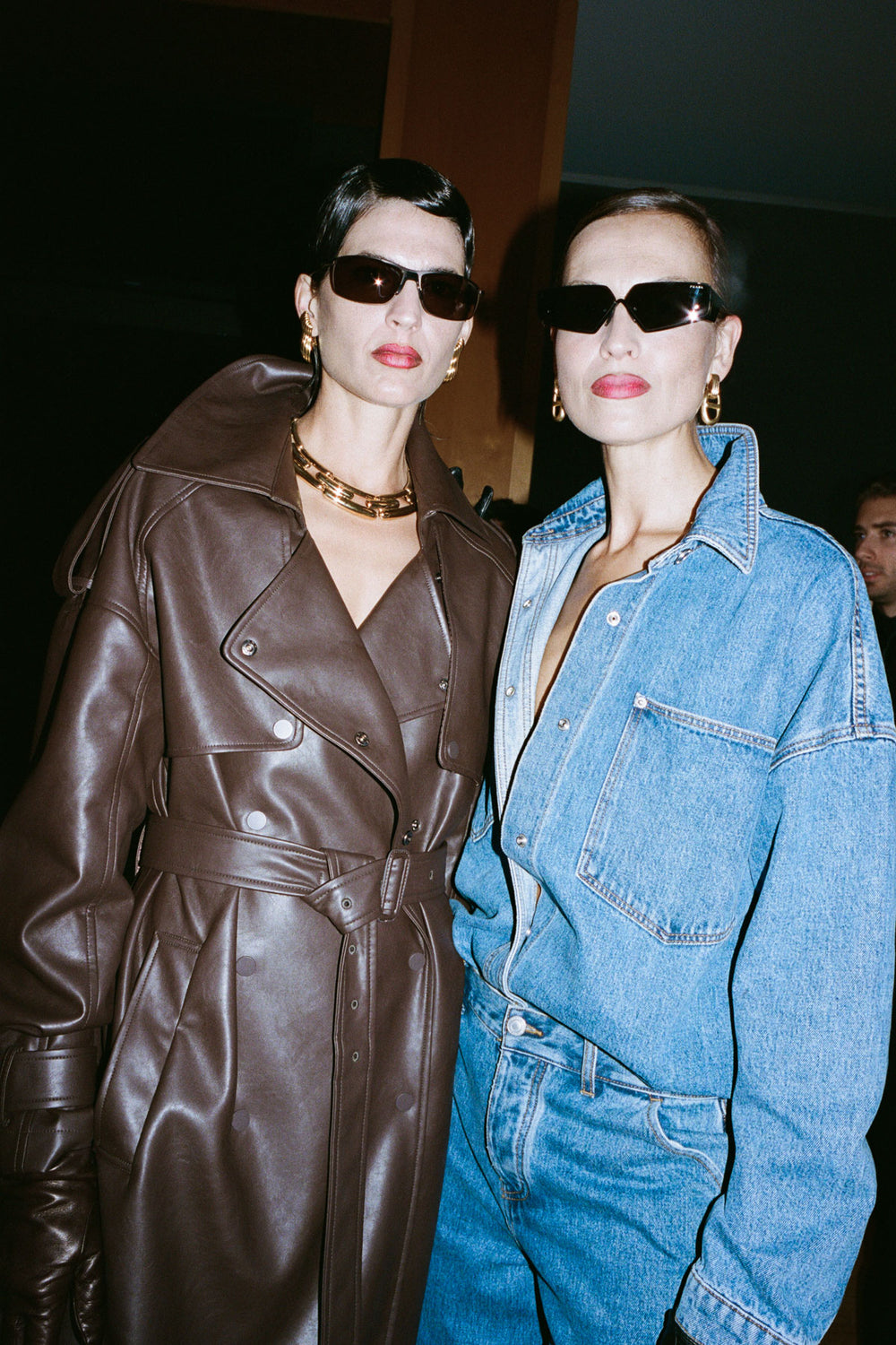 Two fashion models posing together. The model on the left wears a dark brown leather trench coat and sunglasses, while the model on the right wears a light blue denim jacket and jeans with black sunglasses.