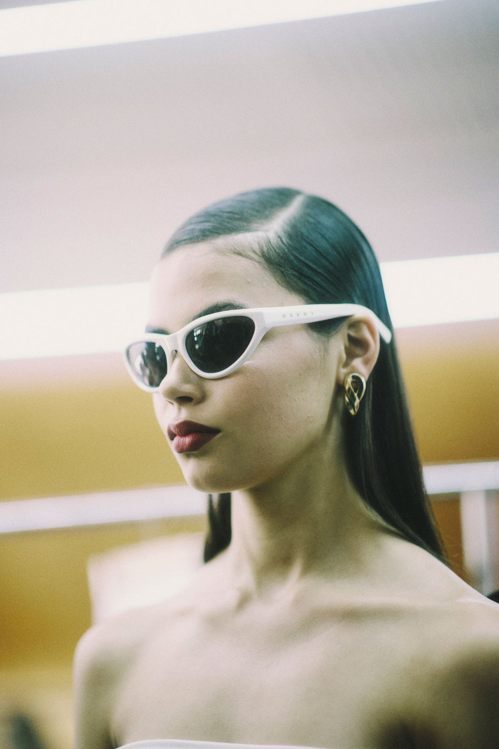 A woman with dark hair pulled back wears white cat-eye sunglasses with dark lenses, shown in a close-up portrait. She has red lipstick on and a small gold hoop earring, with a softly blurred indoor background.