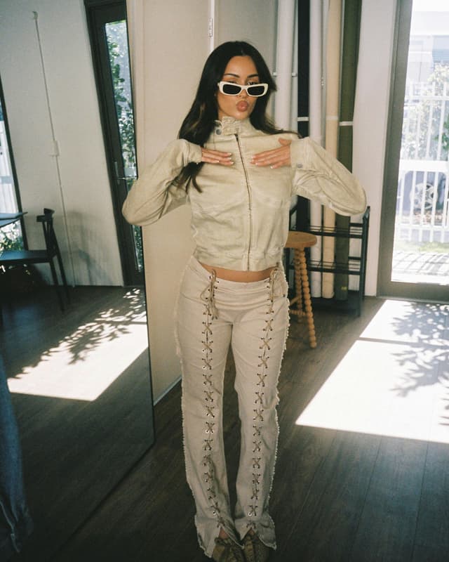 A woman modeling a matching cream-colored two-piece outfit, styled with white-framed jacquemus sunglasses in a sunlit room.
