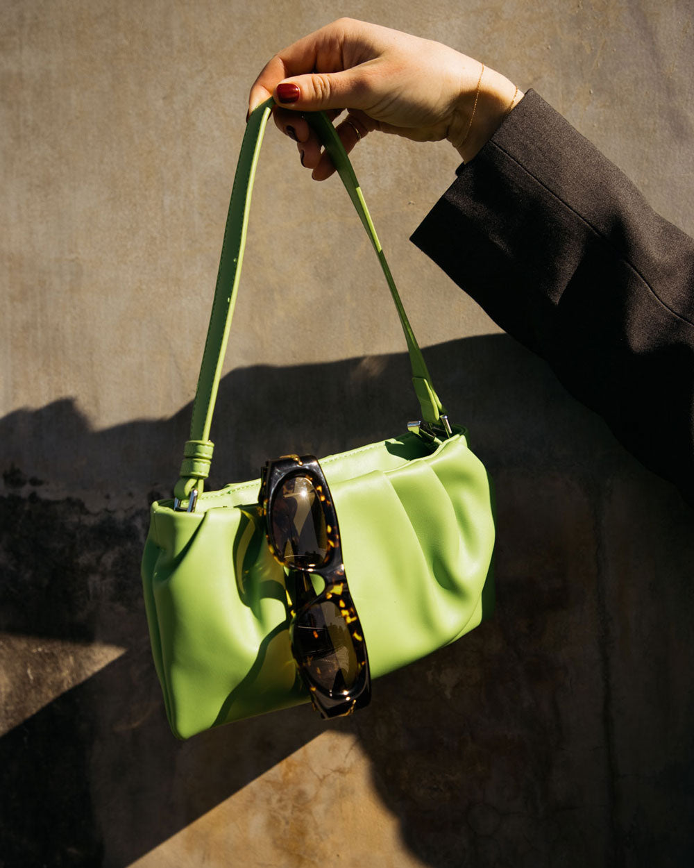 A lime green leather shoulder bag with a matching strap, held by a hand wearing a black sleeve. A pair of tortoiseshell sunglasses is draped over the front of the bag, set against a textured concrete wall with dramatic sunlight and shadows.