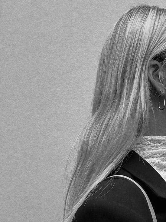A black and white side-profile portrait of a woman with long platinum blonde hair wearing dark sunglasses with 'mi mi mi mi mi mi' printed on the temples, a textured high-neck top, and a dark blazer against a plain light background.