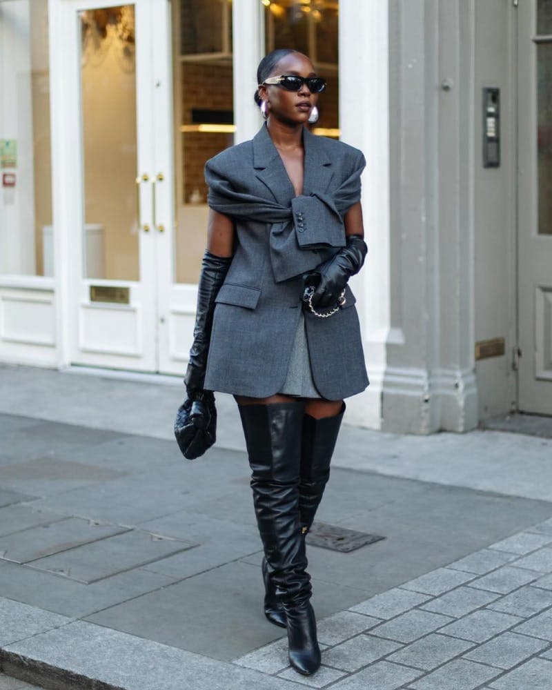 A woman walking on a city sidewalk wearing a gray oversized blazer with a large bow detail, black gloves, black knee-high boots, and black sunglasses.