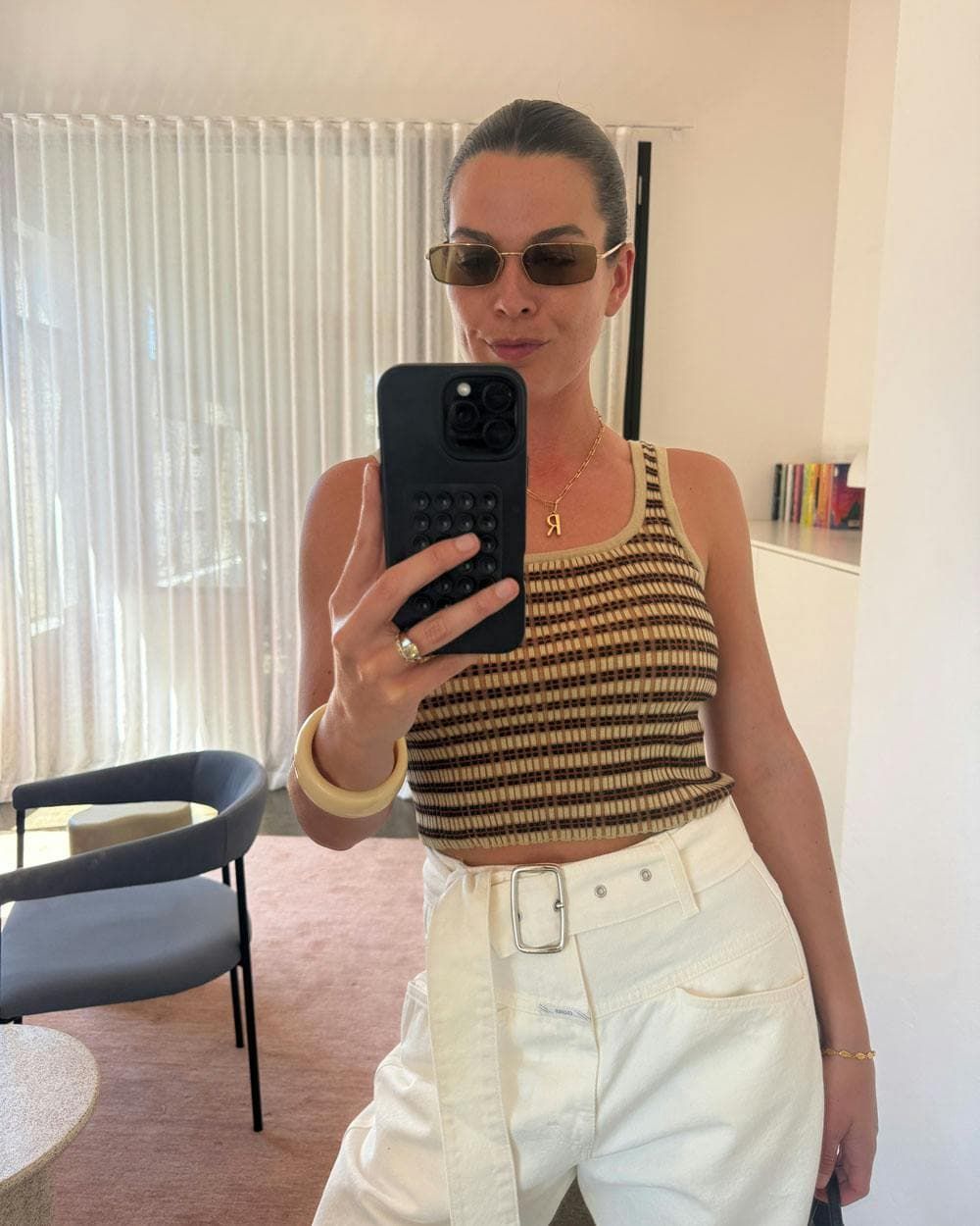Rhiannon joyce taking a mirror selfie in a room, wearing a brown and beige striped knit crop top, white pants, and prada sunglasses.