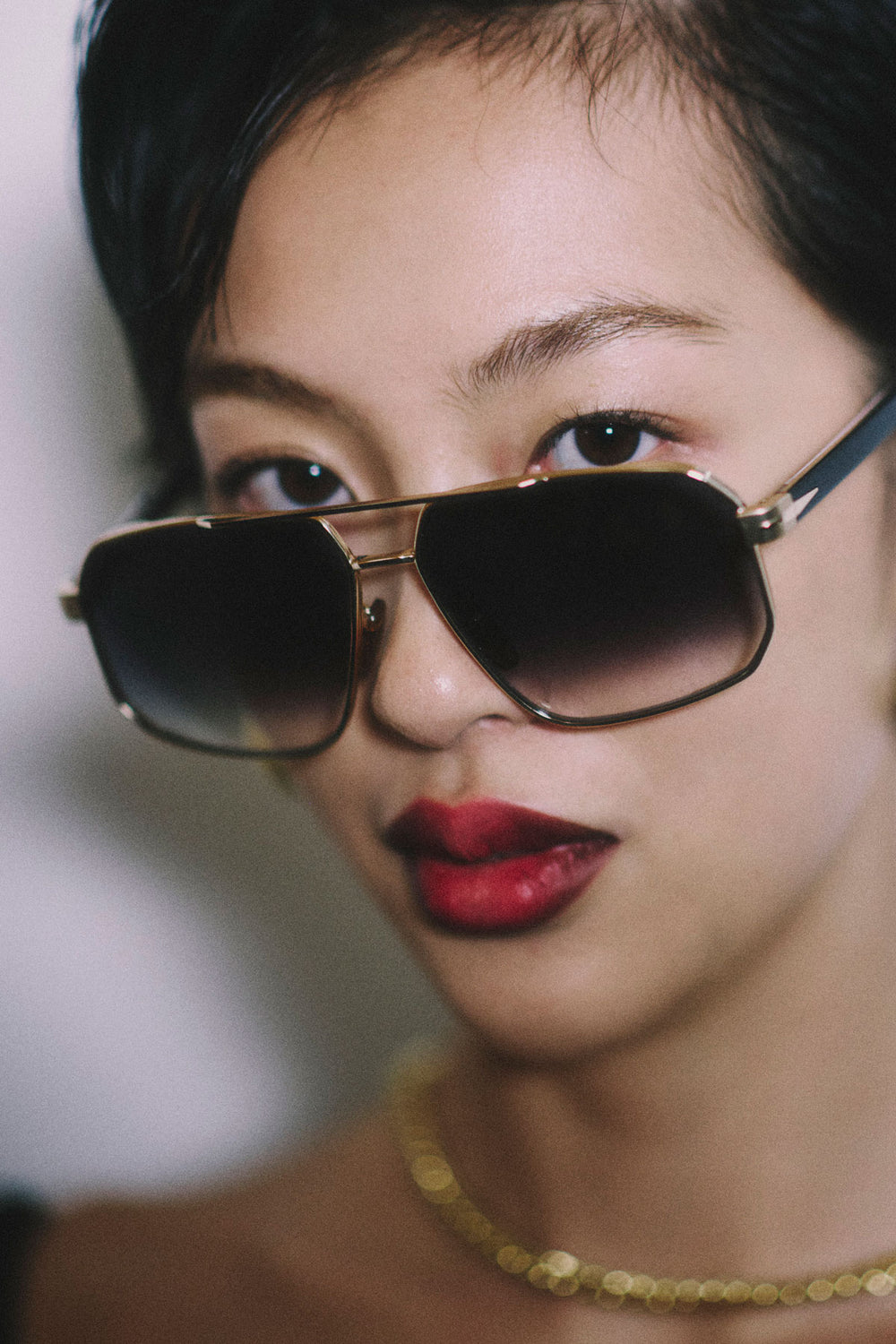 Close-up of a woman wearing oversized aviator-style sunglasses with a dark gradient lens and gold-toned metal frame, paired with red lipstick and a gold chain necklace.