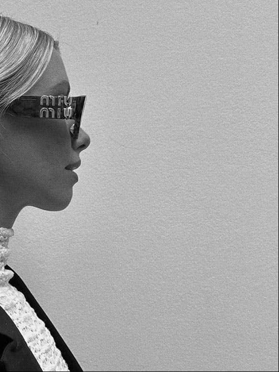 A black and white side-profile portrait of a woman with long platinum blonde hair wearing miu miu sunglasses with 'mi mi mi mi mi mi' printed on the temples, a textured high-neck top, and a dark blazer against a plain light background.