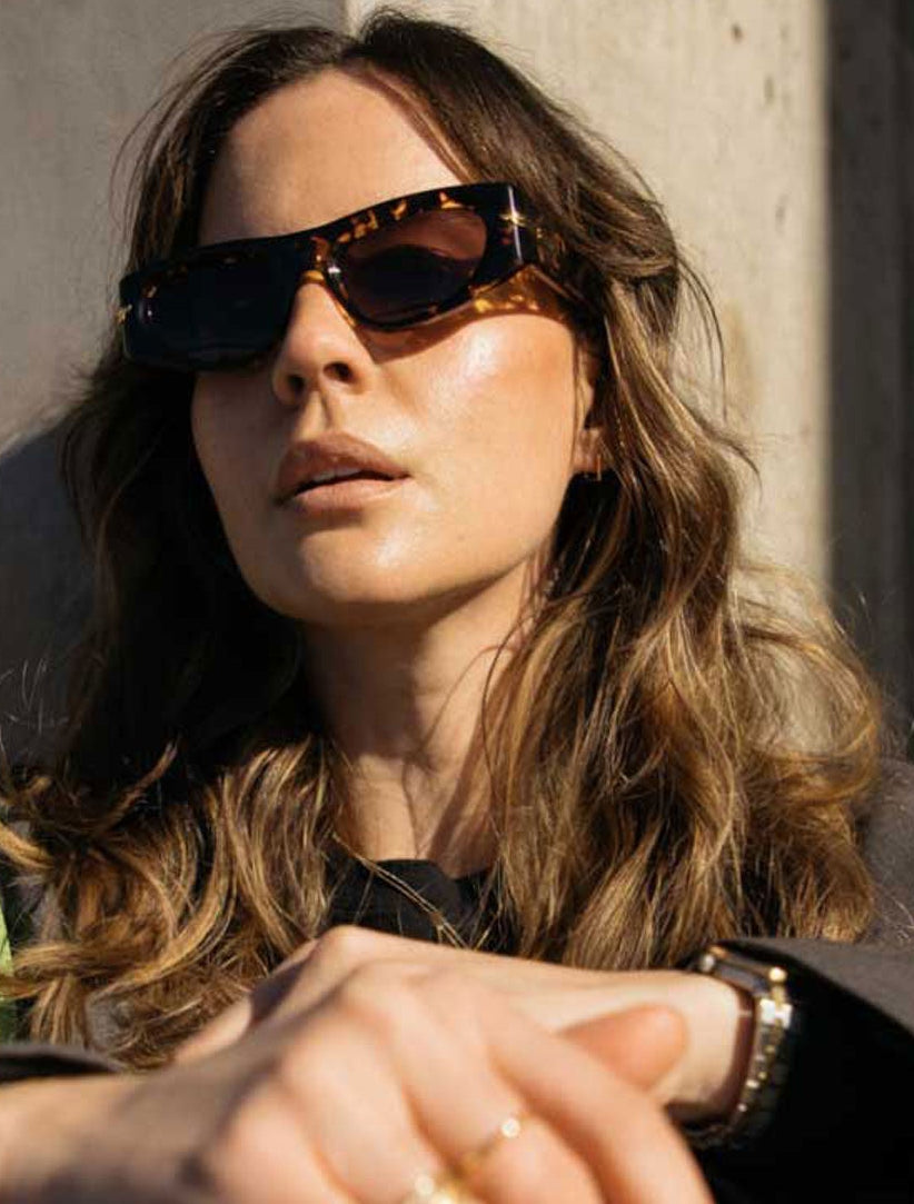 A woman wearing oversized bottega veneta sunglasses with a tortoiseshell pattern and a dark gray blazer, posed against a textured concrete wall in dramatic sunlight.