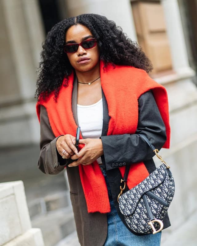 A woman wearing a bright orange-red knit wrap over a black leather jacket, styled with a white top, blue jeans, and a black-and-white Dior monogram shoulder bag, set against a blurred stone building background.