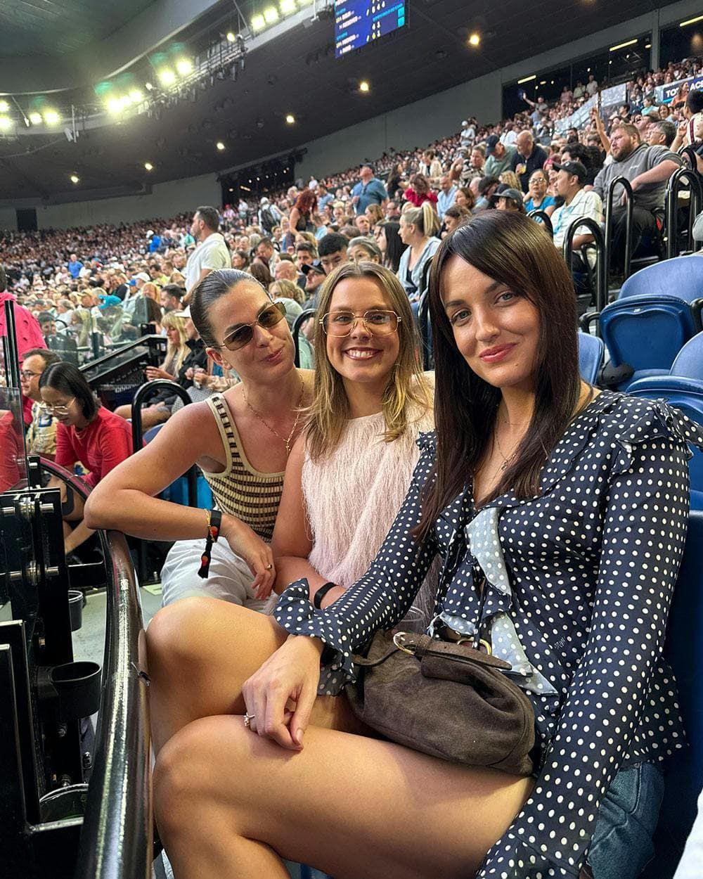 Three women sitting together in crowded stadium seating at an event, with a large audience and arena lights visible in the background.