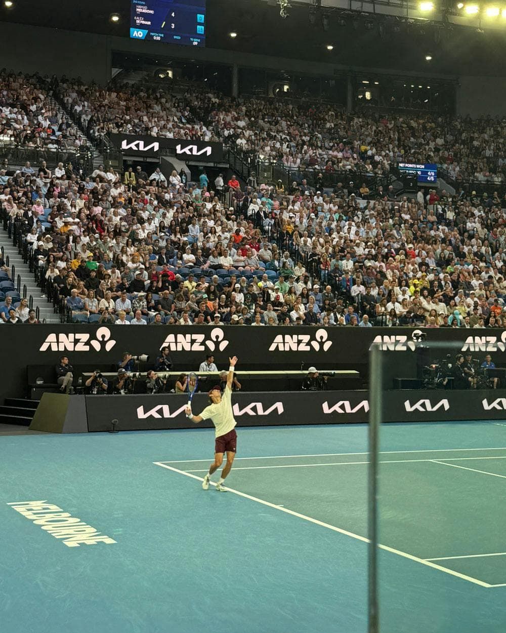 A tennis player serves on a blue indoor court at an arena, with a large crowd in the Australian Open